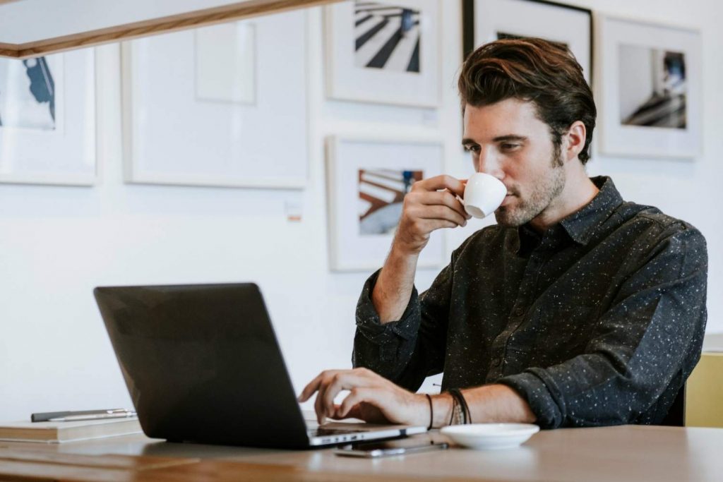 Άντρας πίνει καφέ, πληκτρολογεί στον υπολογιστή του ακουμπώντας σε ξύλινη επιφάνεια γραφείου και ψάχνει θέματα για blogger