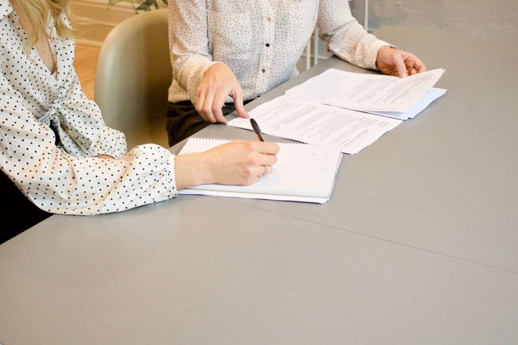 Γυναίκες ελέγχουν βιογραφικό πάνω σε γκρι γραφείο και κρατούν σημειώσεις σε τετράδιο.
