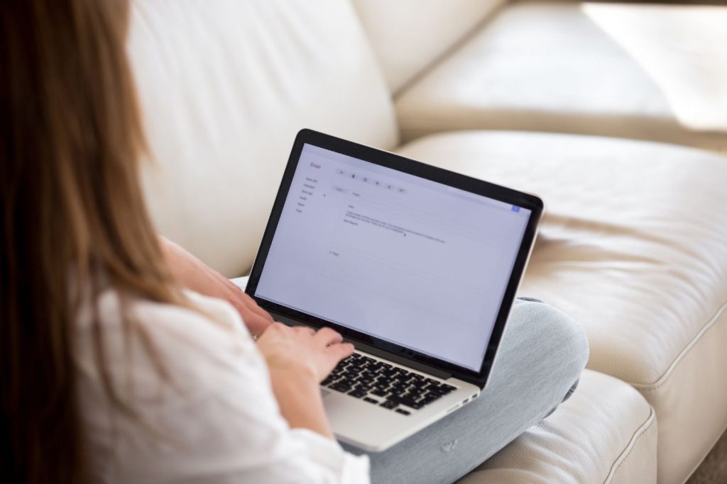 Κοπέλα καθισμένη σε λευκό καναπέ γράφει συνοδευτική επιστολή στο laptop της.