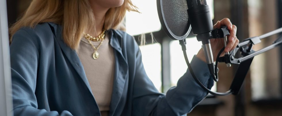 Θεματική εικόνα για: οφέλη voice overs για τις επιχειρήσεις. Νεαρή γυναίκα ηχογραφεί τη φωνή της με επαγγελματικό μικρόφωνο.