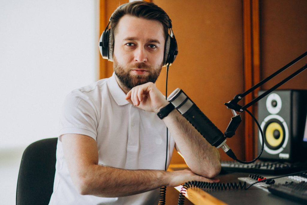 Άντρας μέσα σε στούντιο ηχογράφησης κοιτάει την κάμερα φορώντας ακουστικά και έχοντας μπροστά του ένα επαγγελματικό μικρόφωνο.